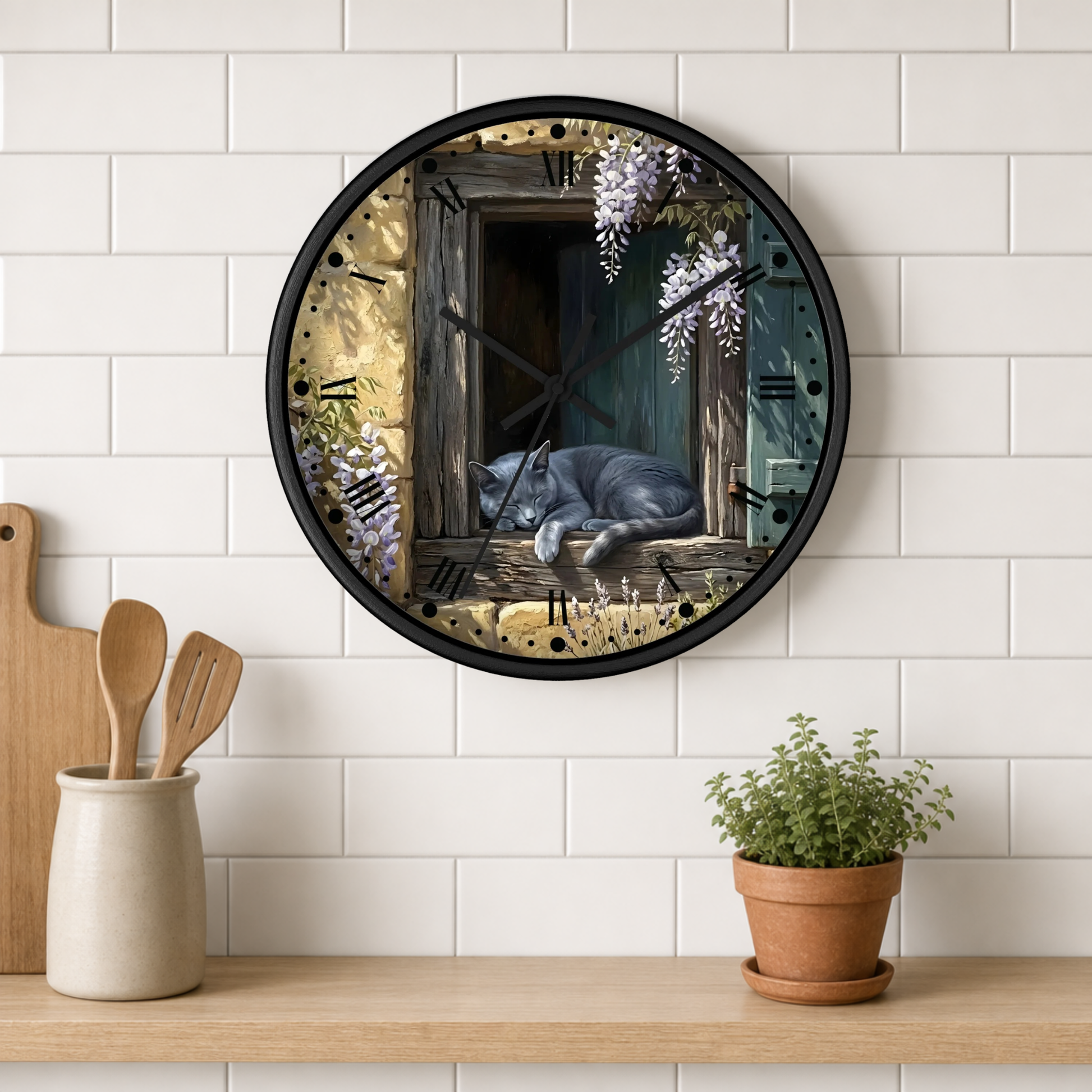 A round wall clock with a black frame and a printed illustration of a gray cat sleeping by an open wooden window with wisteria flowers, mounted on a white subway tile wall above a wooden shelf with kitchen utensils and a potted plant.