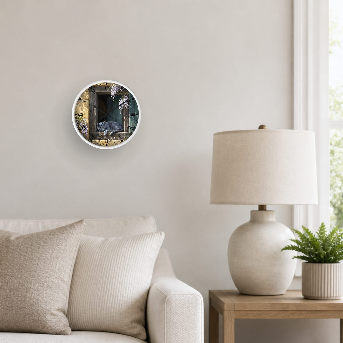 A round wall art print depicting an open window with a gray cat sleeping, styled in a modern living room setting above a beige sofa next to a white table lamp and a small potted plant.