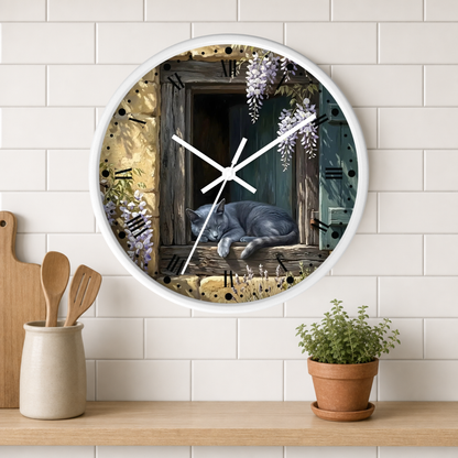 A round wall clock with a white frame hangs on a white subway tile wall above a wooden shelf. The clock face features an illustration of a gray cat sleeping on a rustic wooden windowsill with purple wisteria flowers, a dark green door, and gold-toned background details.