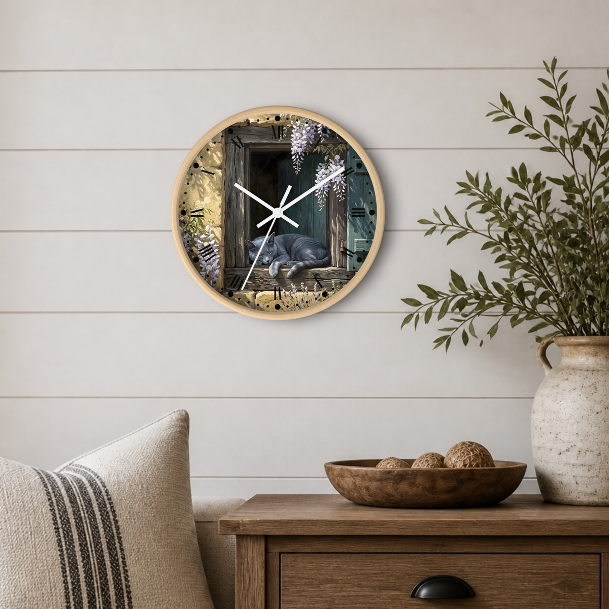 Round wall clock with a light wood frame featuring a printed illustration of a cat in a window, styled on a white shiplap wall above a wooden side table with a plant and decorative bowl.