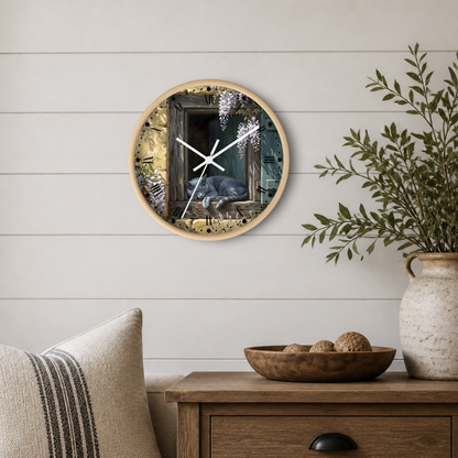 Round wall clock with a light wood frame featuring a printed illustration of a cat in a window, styled on a white shiplap wall above a wooden side table with a plant and decorative bowl.