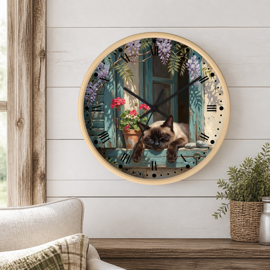 A round wall clock with a light wood frame hangs on a white shiplap wall above a wooden table. The clock face features a printed illustration of a Siamese cat lying on a teal window sill with potted flowers and wisteria.