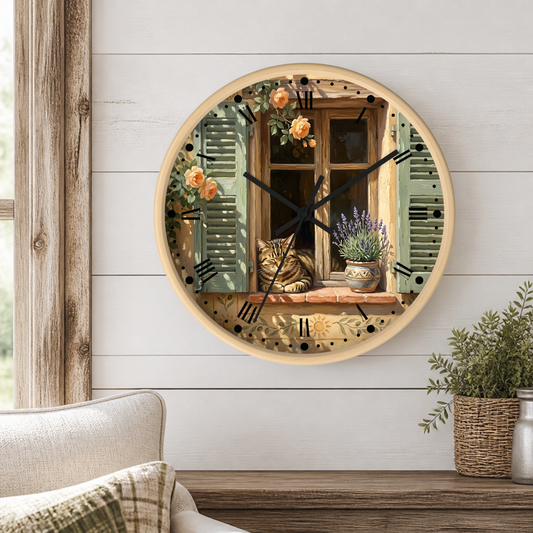 A round wall clock with a rustic French countryside design, featuring a cat on a windowsill with roses and lavender, mounted on a white shiplap wall above a wooden table with a potted plant.