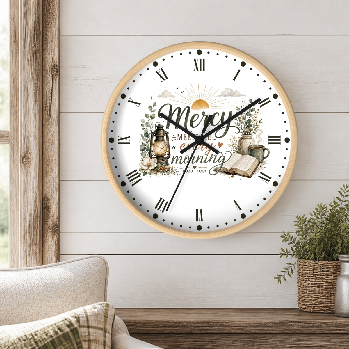 A round wall clock with a light wood frame hangs on a white shiplap wall above a wooden table. The clock face is white with black Roman numerals, black hands, and a decorative center illustration reading 'Mercy in every morning' with a sun, lantern, flowers, and an open book.