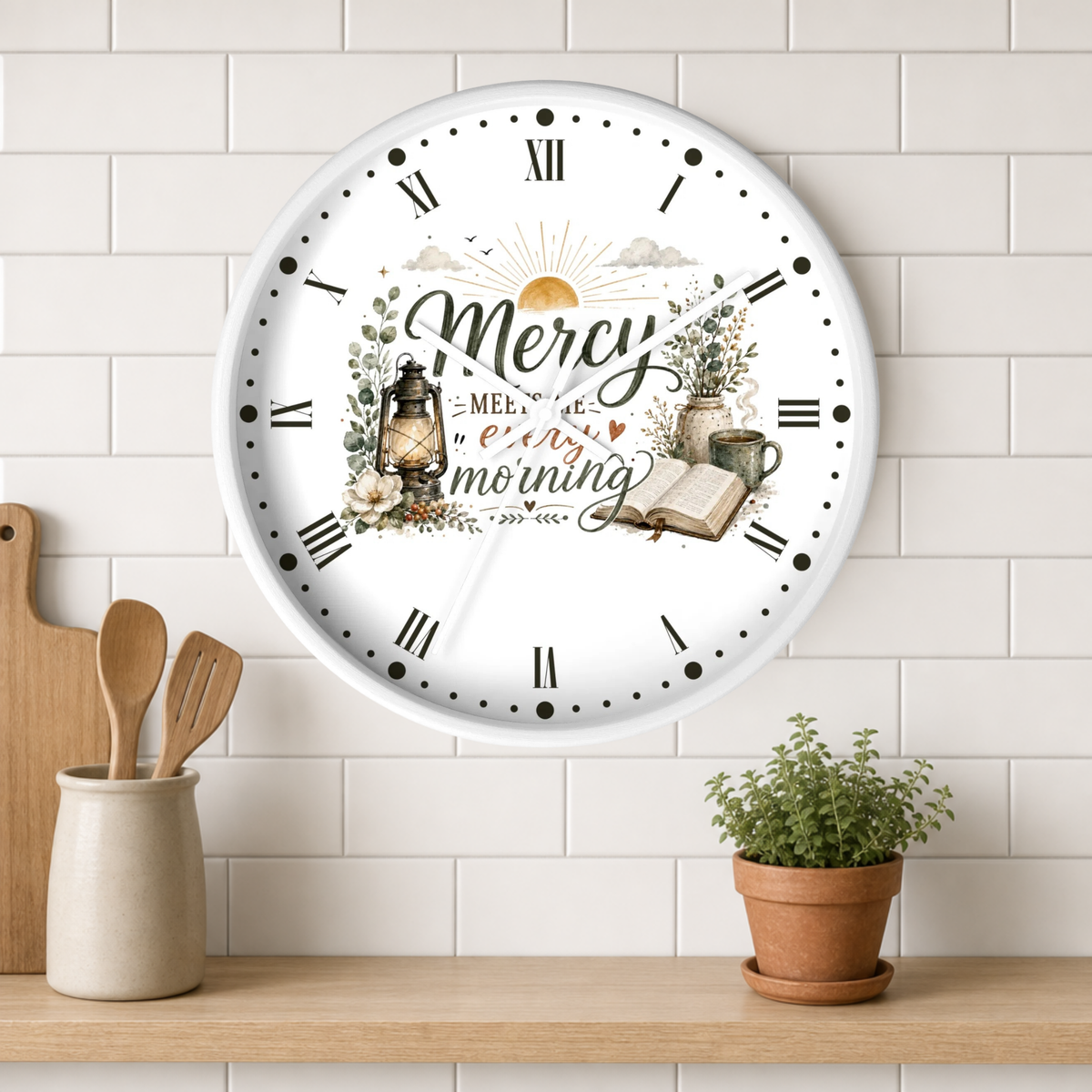 A round wall clock with a white face and Roman numerals, featuring a decorative morning-themed illustration with the words 'Mercy in every morning.' The clock is mounted on a white subway-tile wall above a wooden shelf with a small potted plant and utensils.