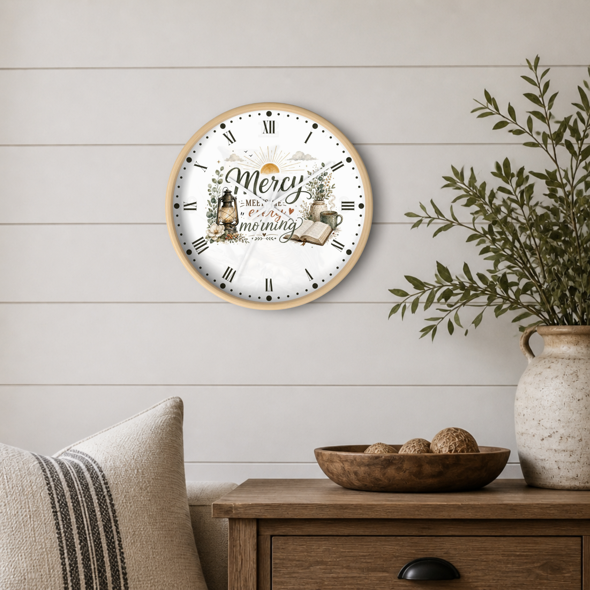 A round wall clock with a light wood frame and a white face featuring the text 'Mercy Meets Me Every Morning' with decorative illustrations, mounted on a white shiplap wall above a wooden side table with a plant and bowl.