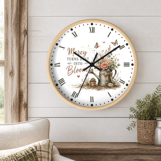 A round wall clock with a light wood frame hangs on a white shiplap wall above a wooden table. The clock face is white with black Roman numerals, black hands, and a central floral illustration featuring a watering can, roses, butterflies, and the words 'Mercy Turns Dust Into Bloom.