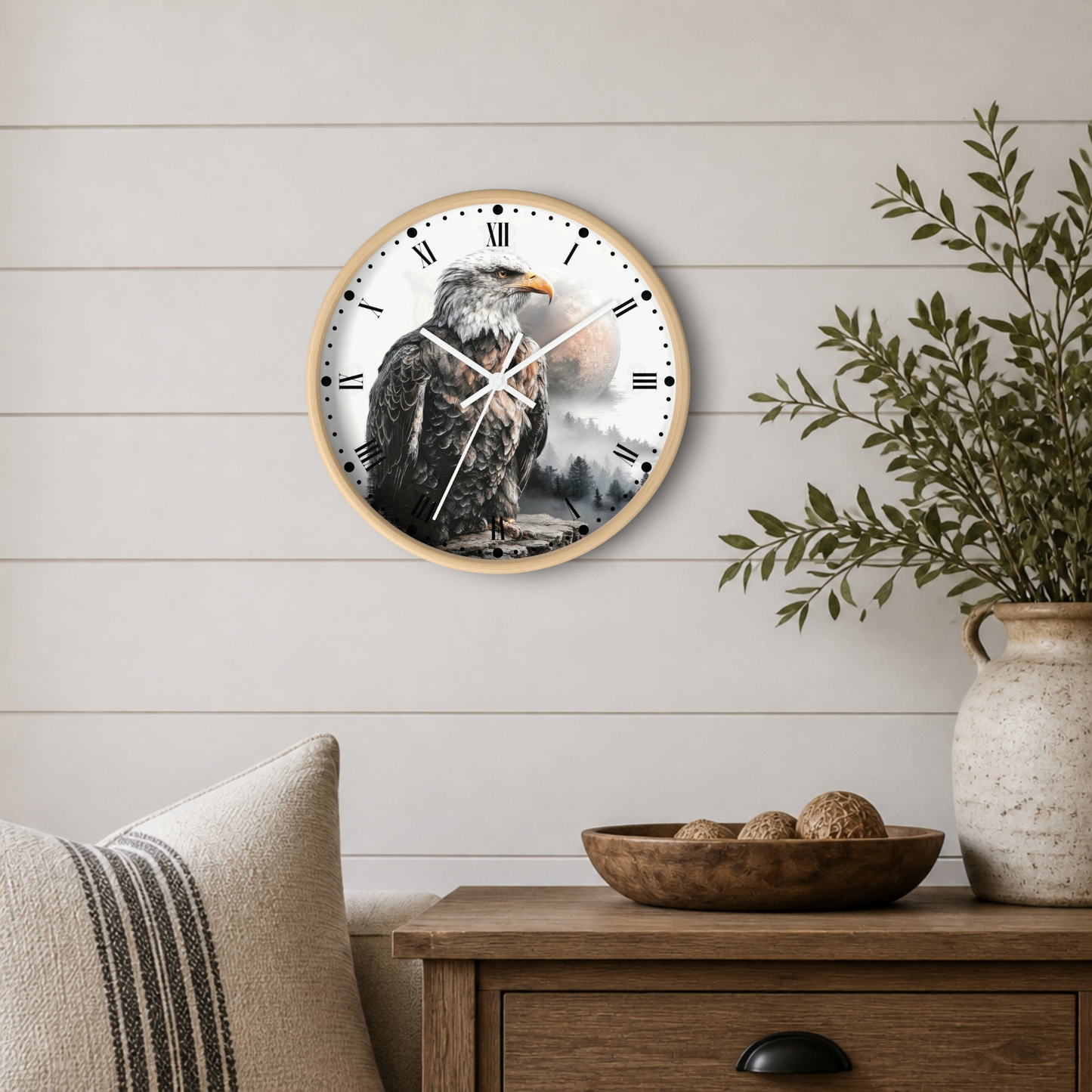 A round wall clock with a light wood frame and a printed eagle design on the face, mounted on a white shiplap wall above a wooden side table with a plant and bowl.
