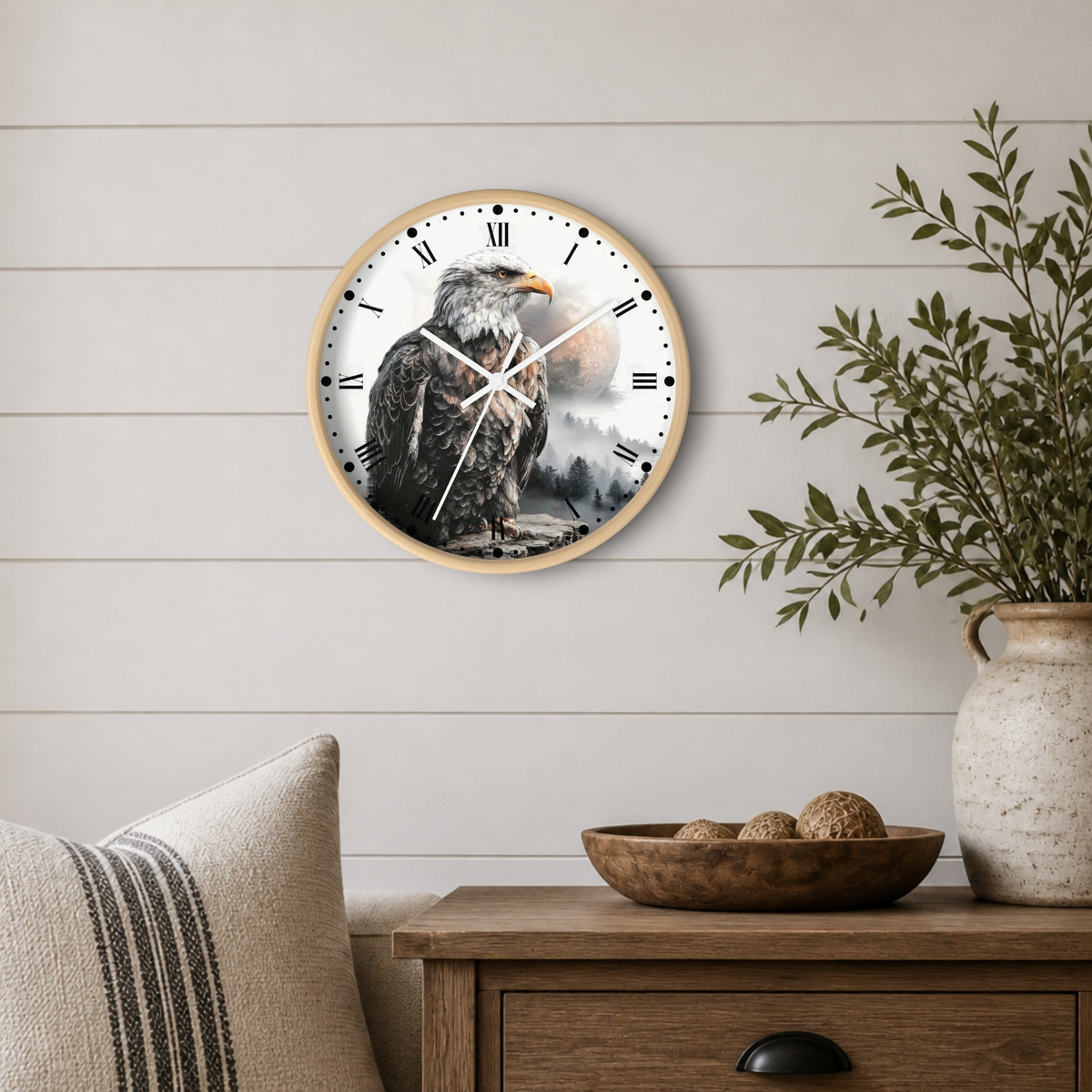 A round wall clock with a light wood frame and a printed eagle design on the face, mounted on a white shiplap wall above a wooden side table with a plant and bowl.