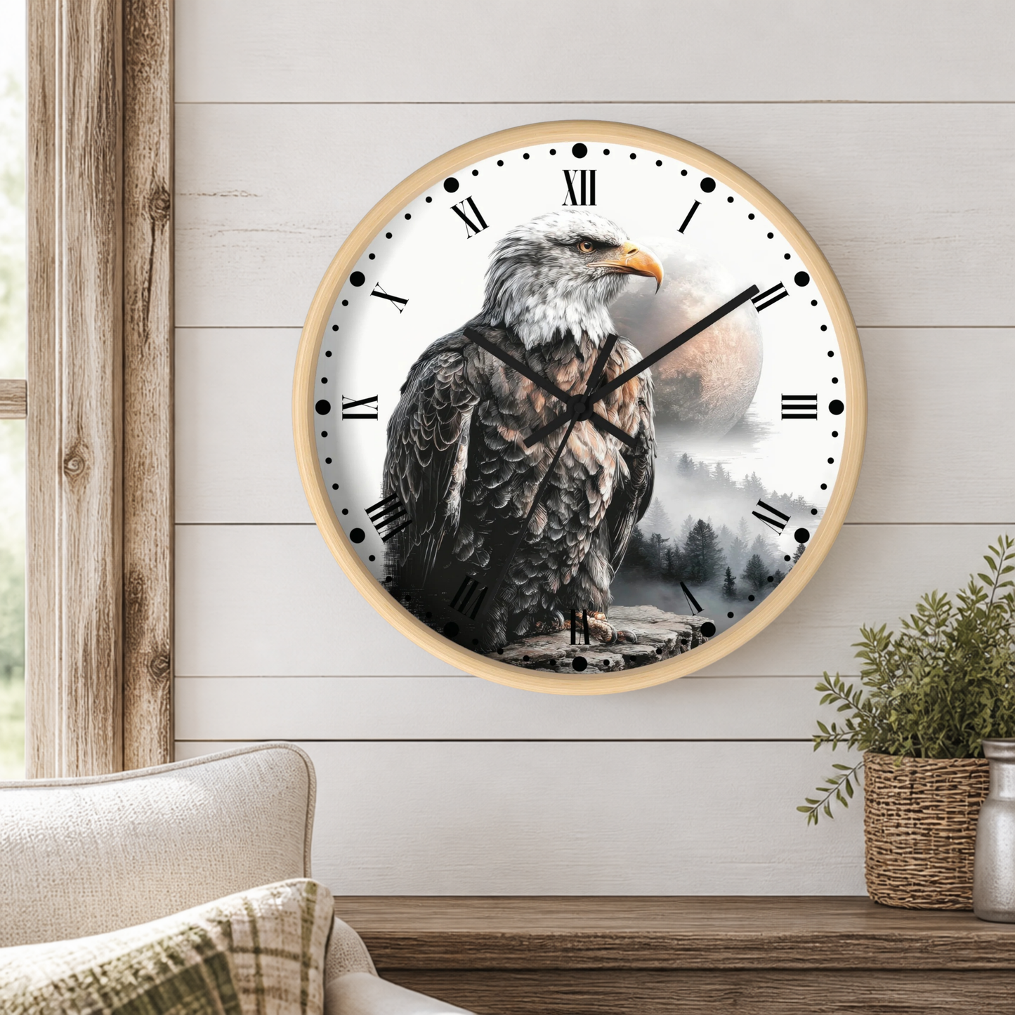 A round wall clock with a light wood frame hangs on a white shiplap wall above a wooden table. The clock face features a detailed black and white illustration of an eagle with a yellow beak, set against a misty mountain landscape with a large moon.