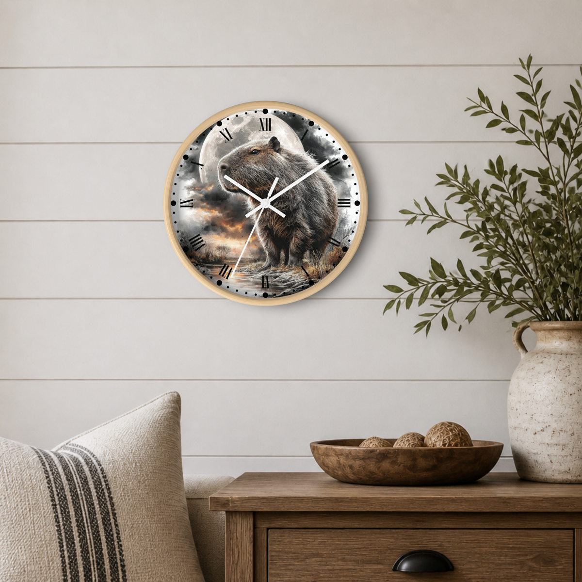 A round wall clock with a natural wood frame hangs on a white shiplap wall above a wooden side table. The clock face features a detailed illustration of a brown capybara standing in front of a full moon, with black Roman numerals and white hands.