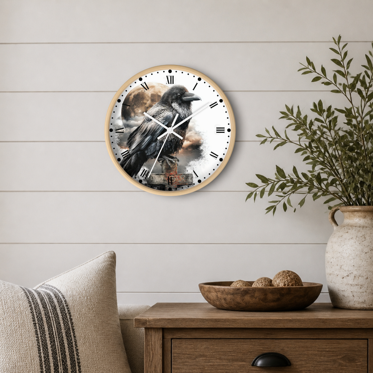 A round wall clock with a light wood frame hangs on a white shiplap wall above a wooden side table. The clock face features a detailed illustration of a raven perched on a branch, with black Roman numerals and clock hands.