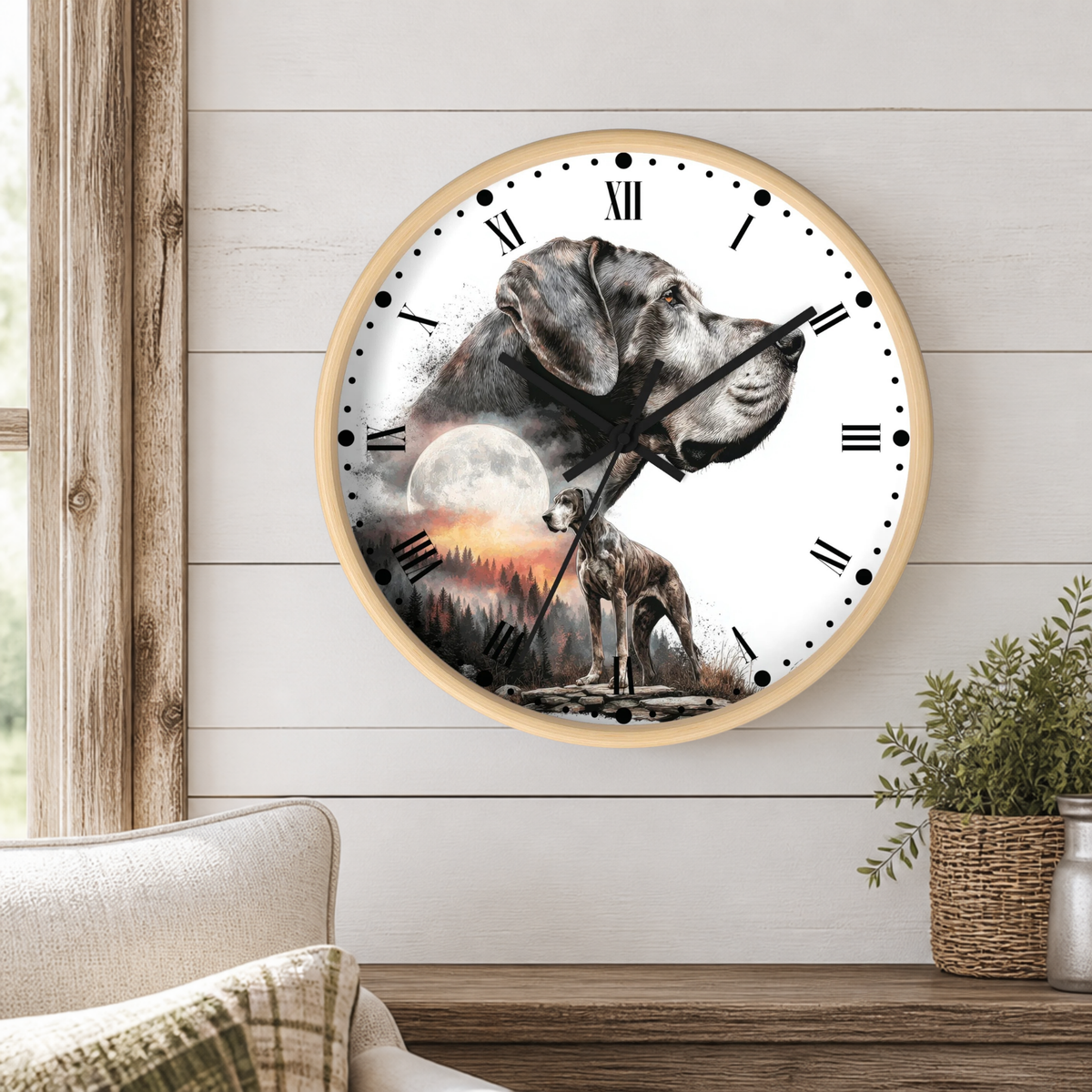 A round wall clock with a light wood frame hangs on a white shiplap wall in a styled room. The clock face features a custom graphic of a gray dog and a smaller dog in a sunset landscape with a full moon.