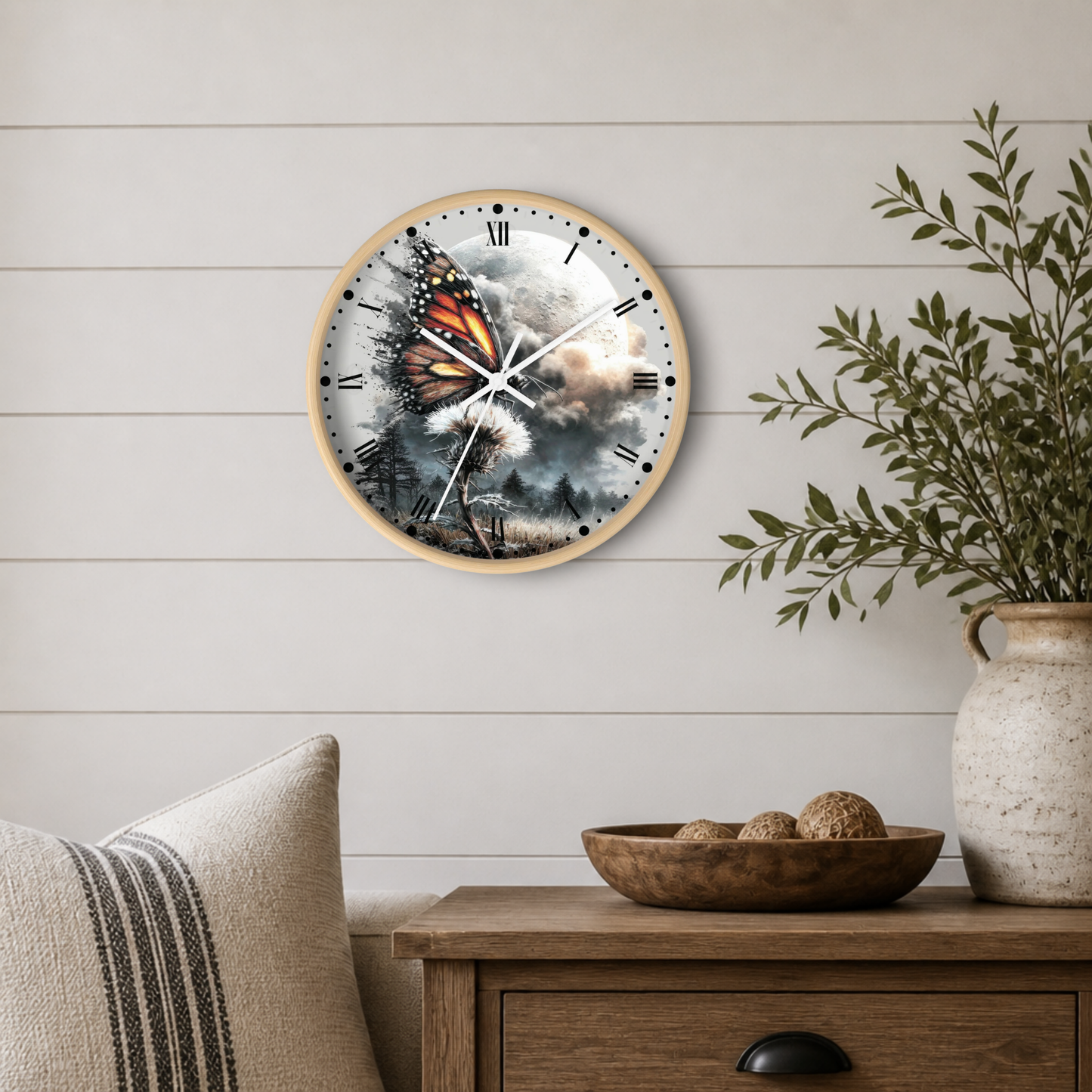 Round wall clock with a light wood frame and a printed butterfly-and-cloud design, mounted on a white shiplap wall above a wooden side table with a plant and decorative bowl.