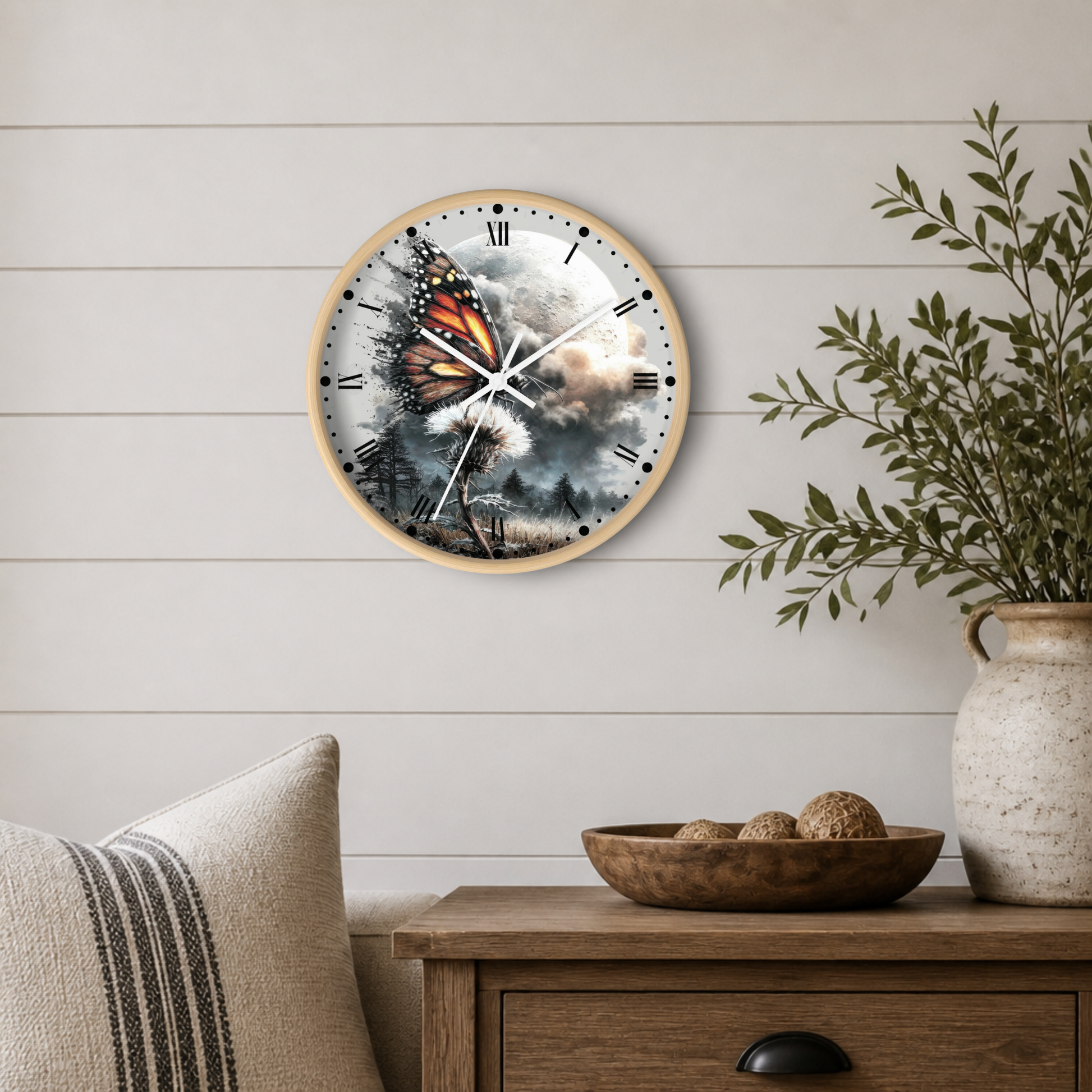 Round wall clock with a light wood frame and a printed butterfly-and-cloud design, mounted on a white shiplap wall above a wooden side table with a plant and decorative bowl.