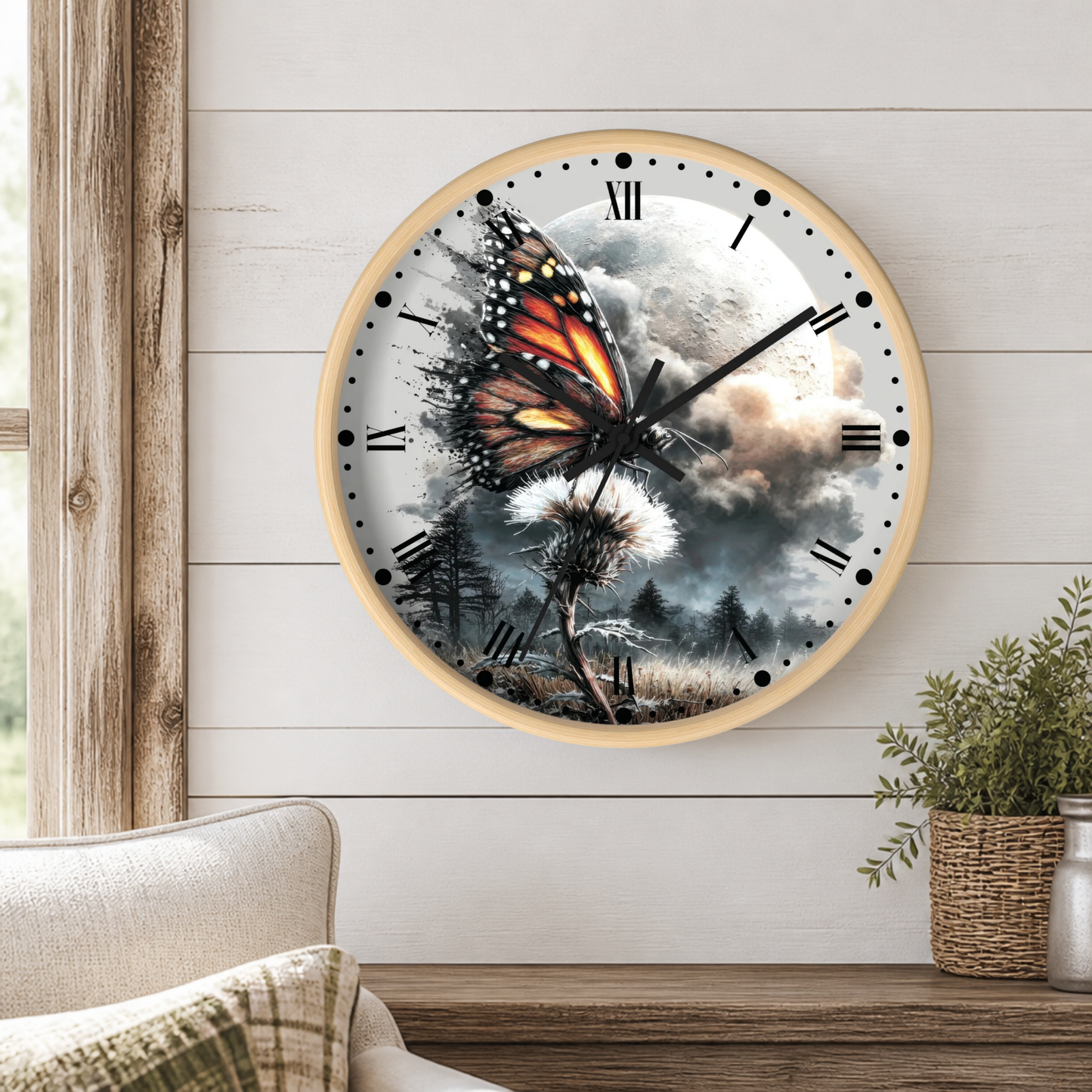 A round wall clock with a light wood frame hangs on a white shiplap wall above a wooden table. The clock face features a surreal illustration of a monarch butterfly emerging from a dandelion, set against a dramatic cloudy sky and moon.