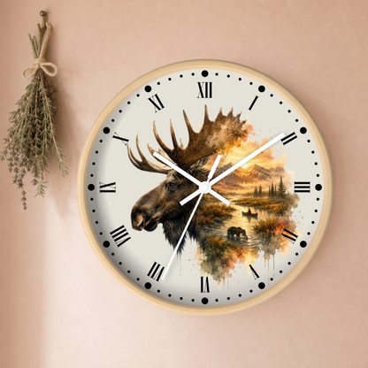 A round wall clock with a light wood frame and a cream dial, featuring a moose head over a scenic landscape illustration. The clock is mounted on a warm beige wall with a dried herb sprig hanging to the left.