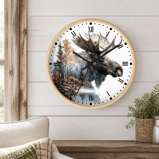 A round wall clock with a light wood frame hangs on a white shiplap wall above a wooden table. The clock face features a detailed illustration of a moose head over a forest and lake scene.