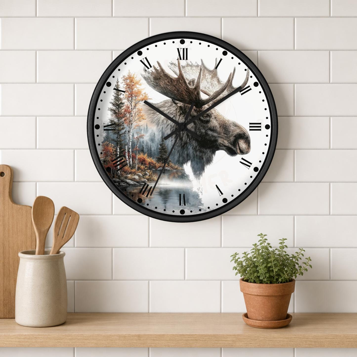 A round wall clock with a black frame and a printed moose head over a forest and lake scene, mounted on a white subway-tile wall above a wooden shelf with kitchen utensils and a potted plant.