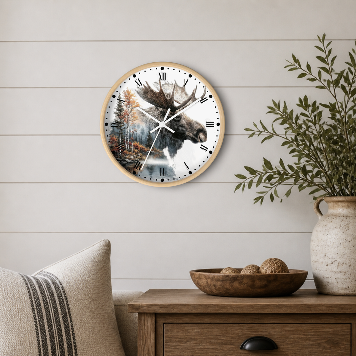 A round wall clock with a light wood frame hangs on a white shiplap wall above a wooden table. The clock face features a detailed illustration of a moose head over a forest and lake scene.