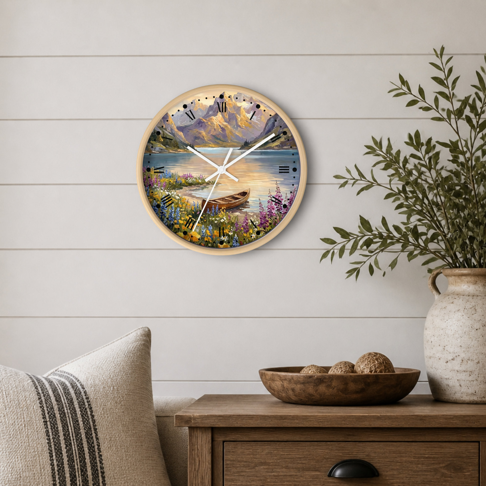 A round wall clock with a natural wood frame and a scenic landscape print of a mountain lake and wildflower meadow, styled on a white shiplap wall above a wooden side table with a vase and bowl.