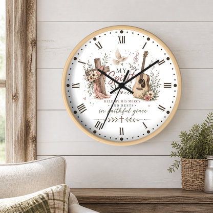 A round wall clock with a light wood frame hangs on a white shiplap wall above a wooden table. The clock face is white with black Roman numerals, black hands, and a central printed design featuring a pink dress, wooden cross, guitar, flowers, a dove, and the words 'My Spirit Sings' and 'Hallelujah His Mercy Rests'.