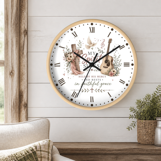 A round wall clock with a light wood frame hangs on a white shiplap wall above a wooden table. The clock face is white with black Roman numerals, black hands, and a central printed design featuring a pink dress, wooden cross, guitar, flowers, a dove, and the words 'My Spirit Sings' and 'Hallelujah His Mercy Rests'.