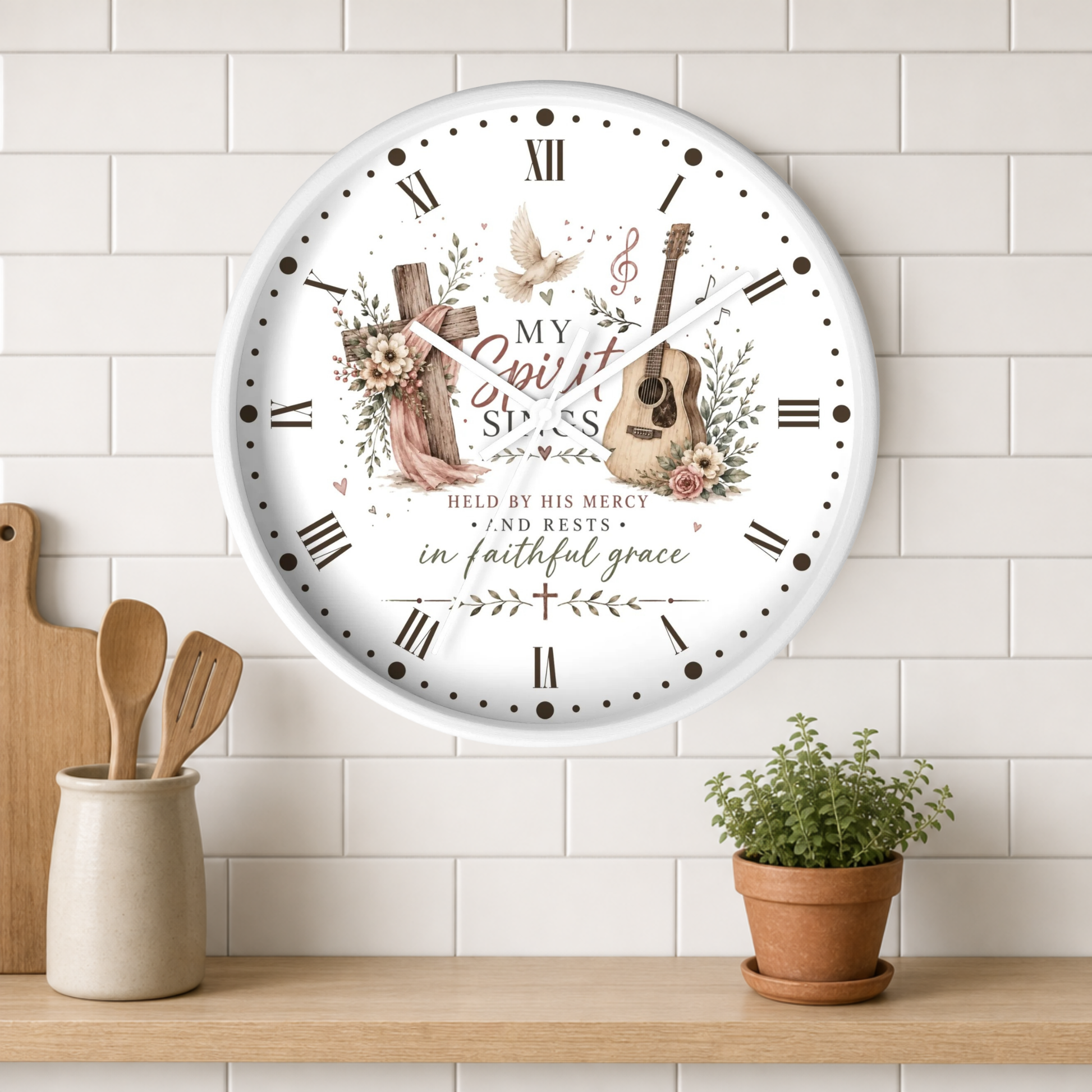 A round wall clock with a white face and black Roman numerals, decorated with a Christian-themed floral and musical illustration including a guitar, cross, dove, and the words 'My Spirit Sings.' The clock is shown hanging on a white subway-tile wall above a wooden shelf with a small potted plant and wooden utensils.