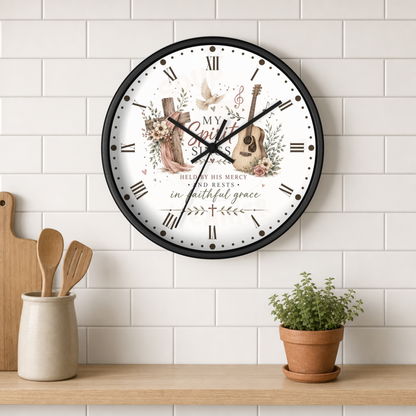 A round wall clock with a black frame and a white face featuring a guitar, piano, floral illustration, and the words 'My God, how precious is the life I may give Him and His mercy and rest.' The clock is mounted on a white subway tile wall above a wooden shelf with kitchen utensils and a small potted plant.