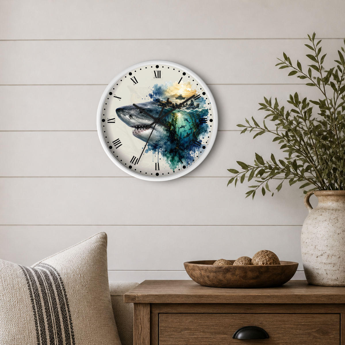 A round wall clock with a white frame and black Roman numerals, featuring a watercolor-style shark illustration in blue, teal, and gray tones. The clock is mounted on a light beige shiplap wall above a wooden side table with a vase of greenery and a pillow in a styled room setting.