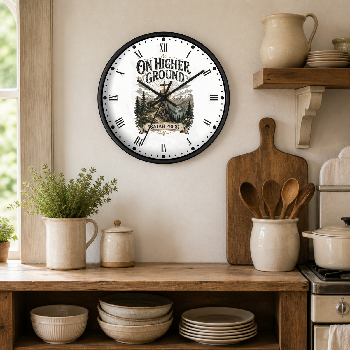 A round wall clock with a black frame and a white clock face featuring the text 'ON HIGHER GROUND' and a mountain illustration, hanging on a beige wall in a rustic kitchen setting with wooden cutting boards, ceramic pitchers, and bowls.