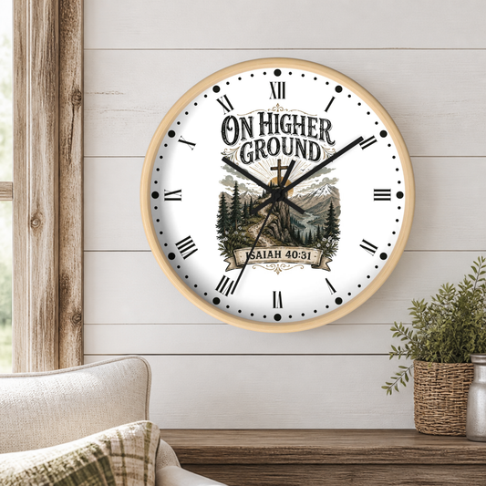 A round wall clock with a light wood frame hangs on a white shiplap wall above a wooden table. The clock face is white with black Roman numerals, black hands, and a scenic mountain-and-tree illustration with the text 'ON HIGHER GROUND' and 'ISAIAH 40:31'.