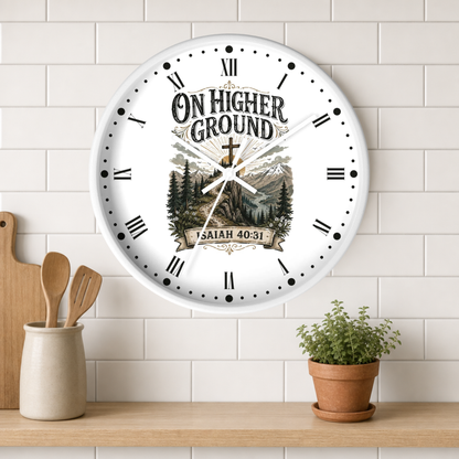 A round white wall clock with Roman numerals and a scenic mountain-and-tree illustration, featuring the text 'ON HIGHER GROUND' and 'ISAIAH 40-31' on the face. The clock is mounted on a white subway tile wall above a wooden shelf with a small potted plant and wooden utensils.