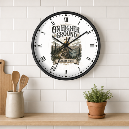 A round wall clock with a black frame and a white face featuring the text 'ON HIGHER GROUND' and 'SAITH 40:31' above a scenic illustration, mounted on a white subway tile wall above a wooden shelf with kitchen utensils and a potted plant.