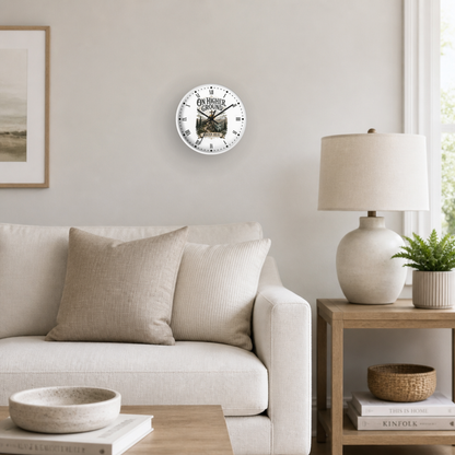 A round wall clock featuring a Christian illustration, mounted on a light beige wall in a styled living room setting with a sofa, side table, lamp, and plant.