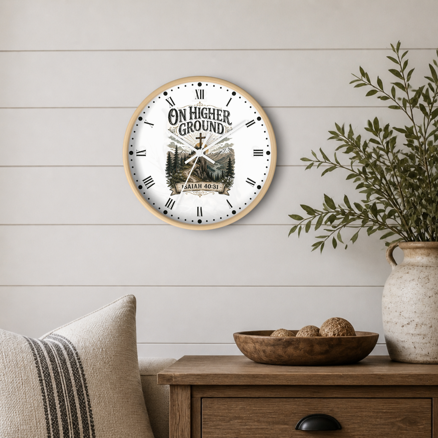 A round wall clock with a light wood frame and a white face featuring the text 'ON HIGHER GROUND' and a mountain-and-cross illustration, styled on a white shiplap wall above a wooden side table with a vase and pillows.