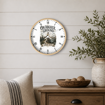 A round wall clock with a light wood frame and a white face featuring the text 'ON HIGHER GROUND' and a mountain-and-cross illustration, styled on a white shiplap wall above a wooden side table with a vase and pillows.