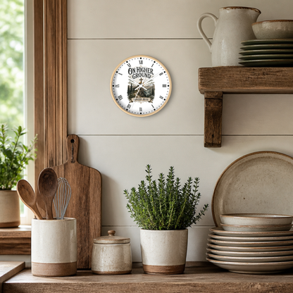 A round wall clock with a light wood frame hangs on a white shiplap wall in a rustic kitchen setting. The clock face is white with black Roman numerals and features a central illustration of a mountain peak above the words 'ON HIGHER GROUND'.