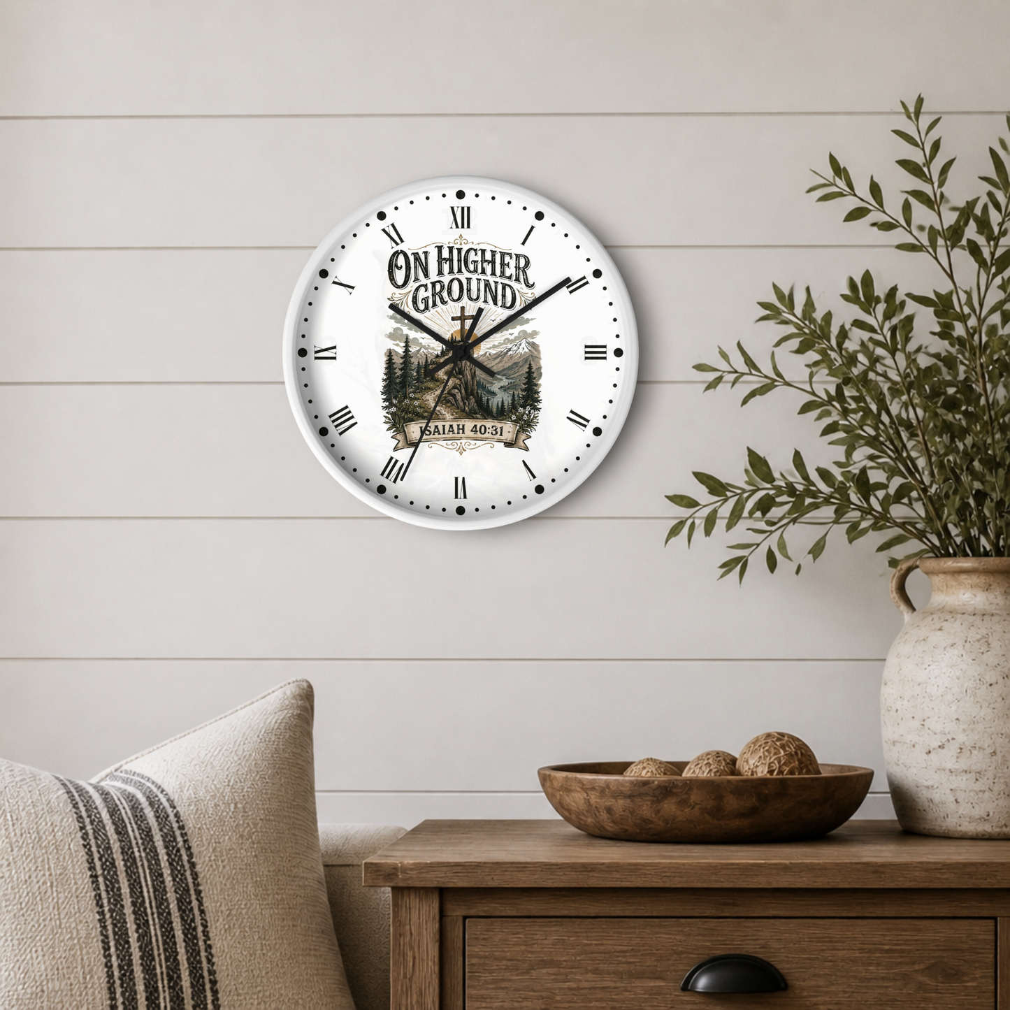 A round wall clock with a white face and black Roman numerals hangs on a light beige shiplap wall above a wooden side table. The clock features a rustic western design with the text 'ON HIGHER GROUND' around a mountain-and-tree illustration.