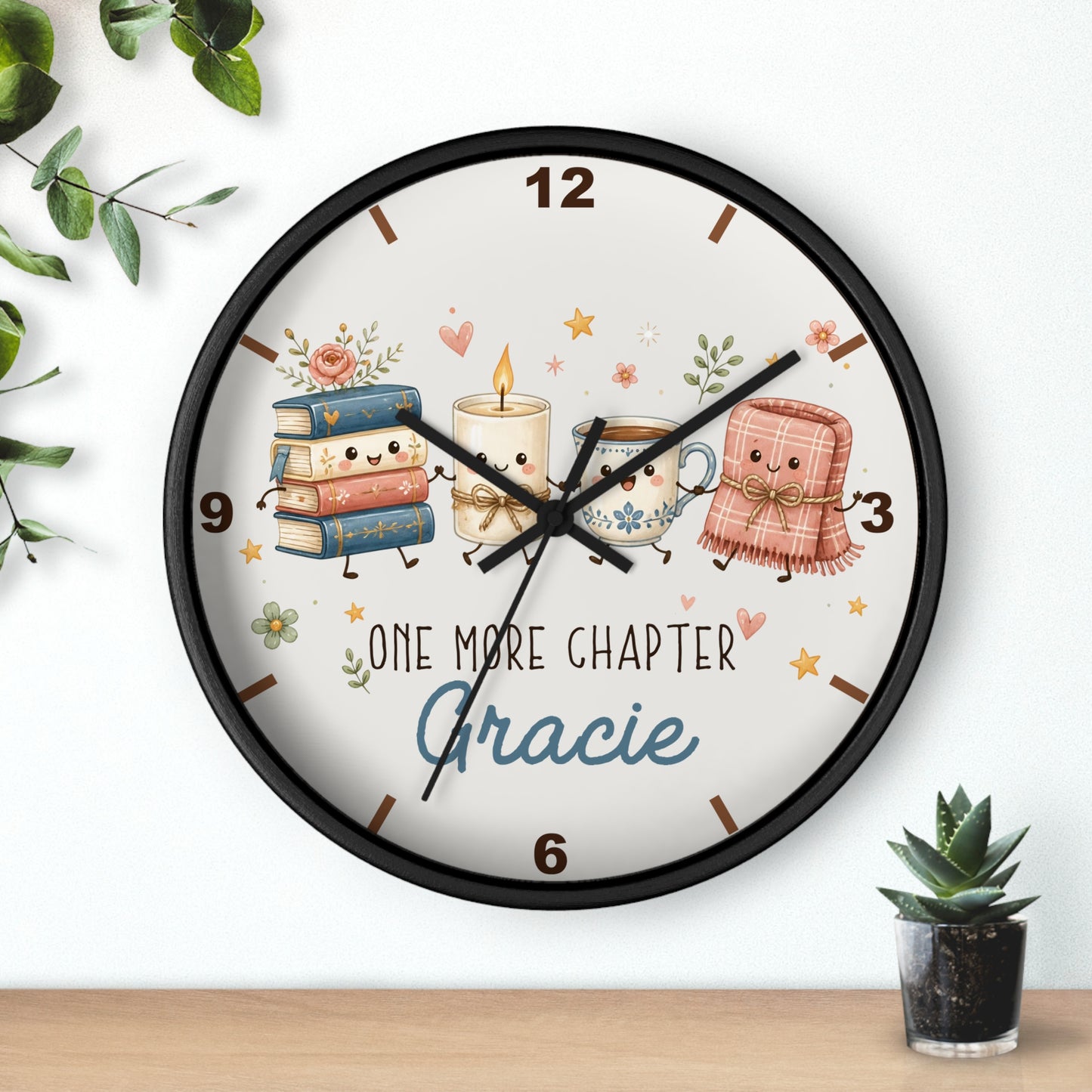A round wall clock with a black frame and a whimsical illustration of books, a candle, a teacup, and a pink blanket character on a cream face. The clock is mounted on a white wall above a light wood surface, with small green plants in the corners.
