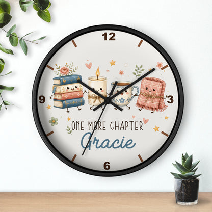 A round wall clock with a black frame and a whimsical illustration of books, a candle, a teacup, and a pink blanket character on a cream face. The clock is mounted on a white wall above a light wood surface, with small green plants in the corners.