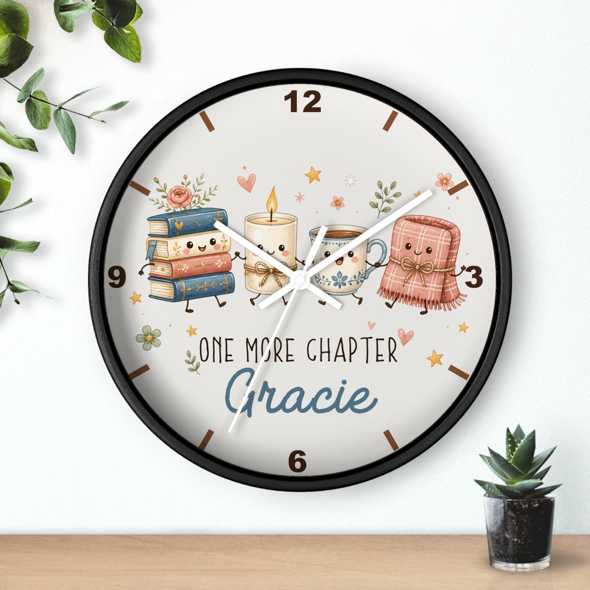 Personalized round wall clock with a black frame and a whimsical illustration of books, a candle, a mug, and a pink blanket with the text 'ONE MORE CHAPTER Gracie' on a white background, styled on a wall above a wooden surface with small plants.