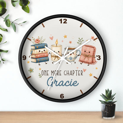 Personalized round wall clock with a black frame and a whimsical illustration of books, a candle, a mug, and a pink blanket with the text 'ONE MORE CHAPTER Gracie' on a white background, styled on a wall above a wooden surface with small plants.