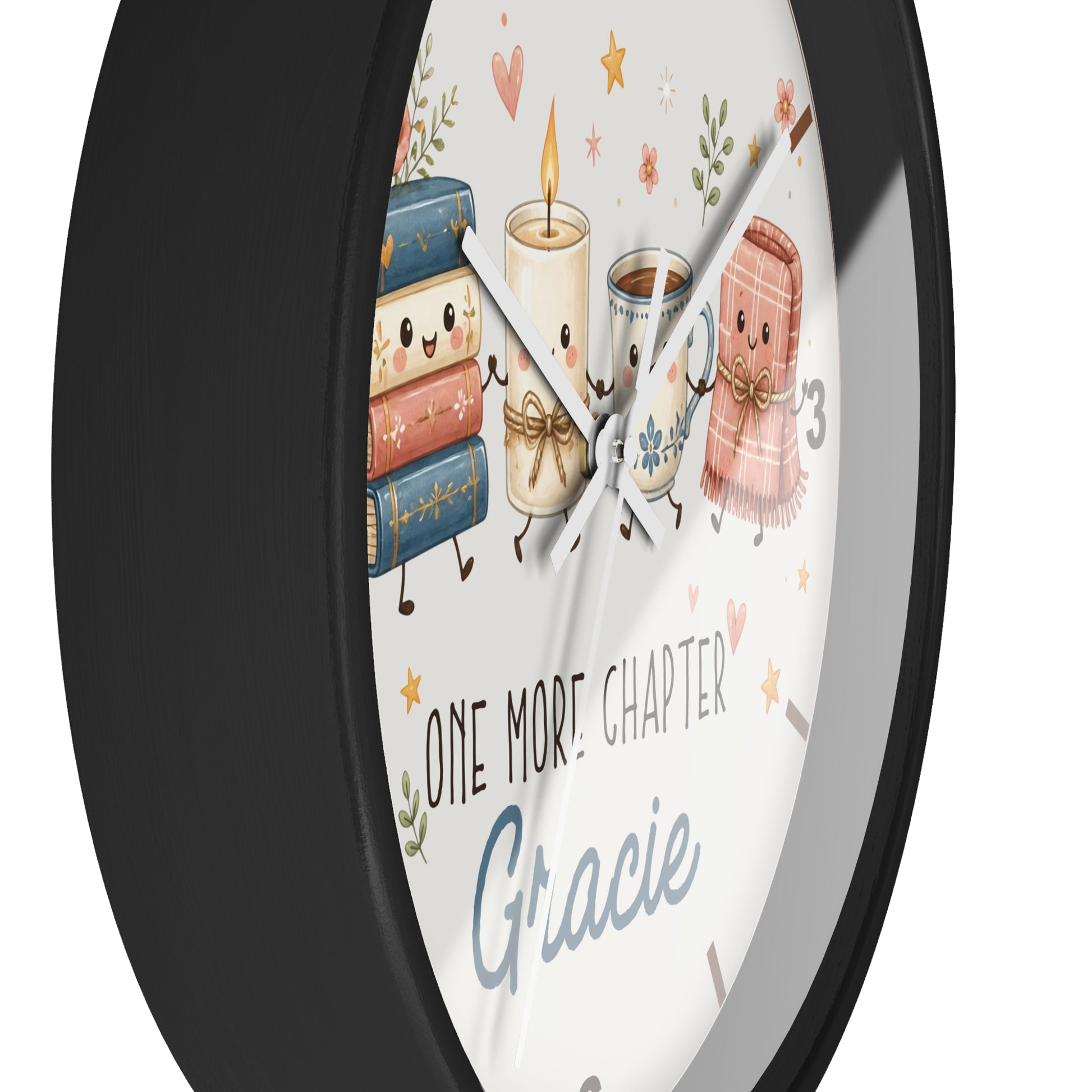 A round wall clock with a black frame and a white face featuring cute illustrated books, a candle, a mug, and a pink blanket. The clock face includes the text 'ONE MORE CHAPTER' and 'Gracie'.
