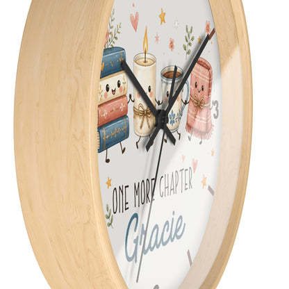 A round wall clock with a light natural wood frame and a white face featuring cute illustrated books, a candle, a mug, a scarf, and the text 'ONE MORE CHAPTER Gracie'.