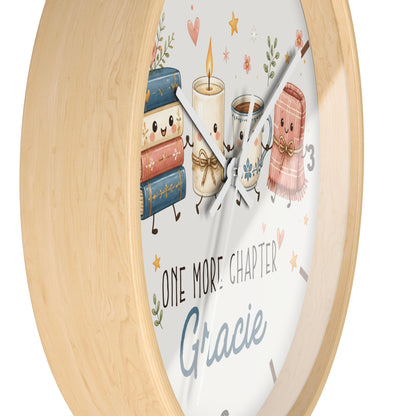 A round wall clock with a light natural wood frame and a white face featuring cute illustrated books, a candle, a mug, and a pink blanket. The clock face includes the text 'ONE MORE CHAPTER' and the name 'Gracie' in blue script.