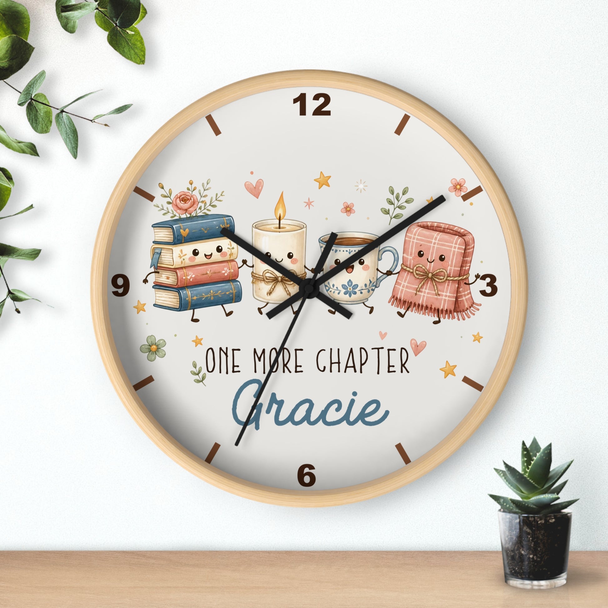 A round wall clock with a light wood frame and a whimsical book-themed dial. The dial features cute illustrated books, candles, and a teacup with the text 'ONE MORE CHAPTER Gracie'.