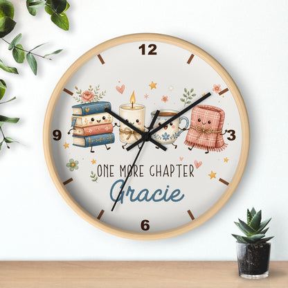 A round wall clock with a light wood frame and a whimsical book-themed dial. The dial features cute illustrated books, candles, and a teacup with the text 'ONE MORE CHAPTER Gracie'.