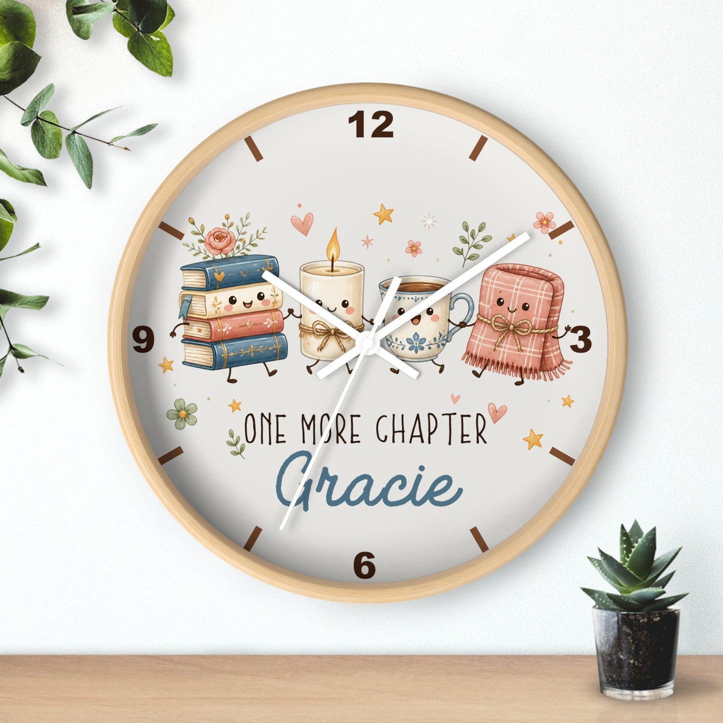 A round wooden wall clock with a light wood frame and a whimsical book-themed dial. The dial features cute illustrations of a stack of books, a candle, a mug, and a pink blanket with faces, alongside the text 'ONE MORE CHAPTER Gracie'.