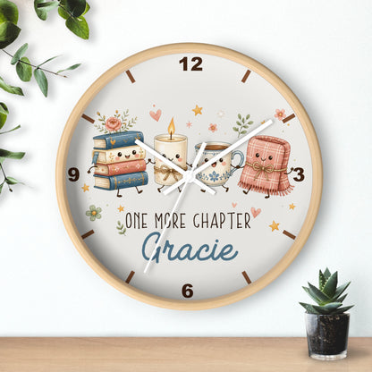 A round wooden wall clock with a light wood frame and a whimsical book-themed dial. The dial features cute illustrations of a stack of books, a candle, a mug, and a pink blanket with faces, alongside the text 'ONE MORE CHAPTER Gracie'.